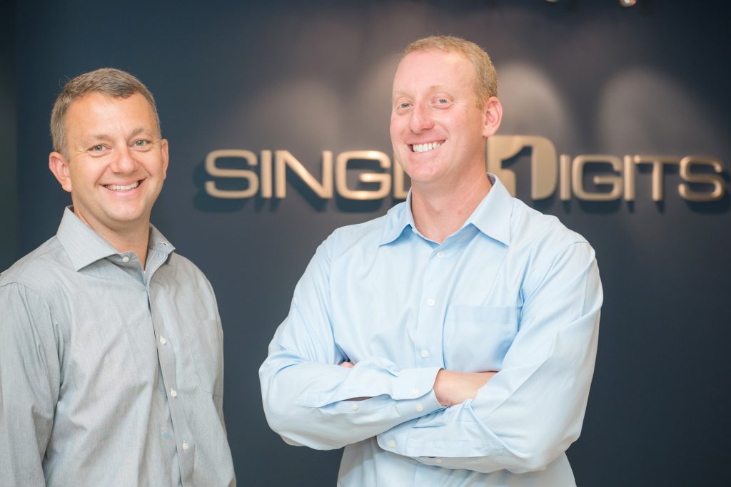 Two men stand in front of the Single Digits logomounted on the wall